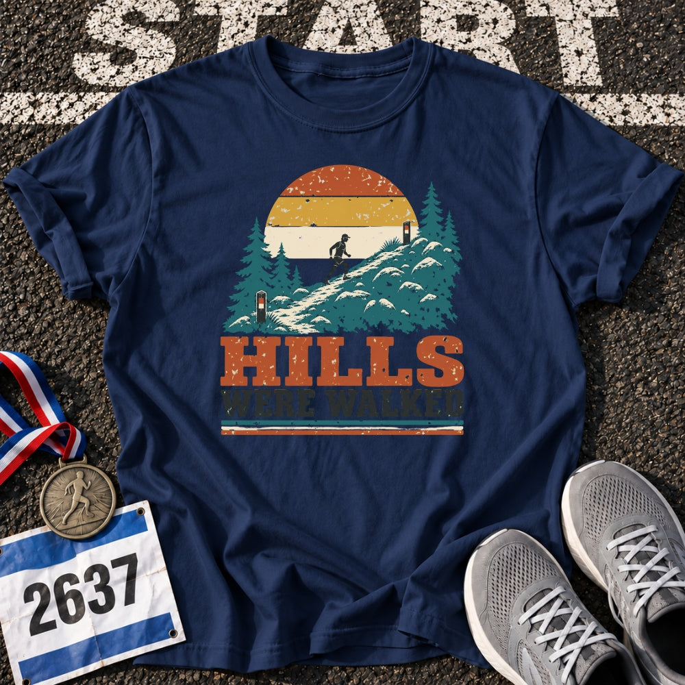 A navy blue graphic t-shirt laid flat on asphalt, featuring a retro sunset mountain scene with pine trees and the text 'HILLS WE'WASHIRE'. Surrounding props include a race medal, race bib number 2637, and gray running shoes.